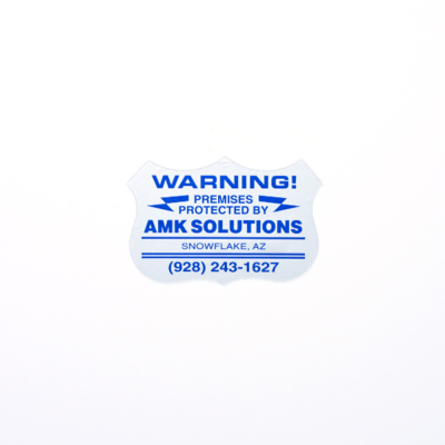 A small badge sticker that's white with blue text that says 'Warning premises protected by  AMK Solutions'