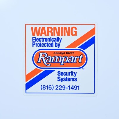 A square sticker that is white with an orange and blue design on it that's a warning label for Rampart security systems